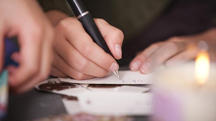 Nahaufnahme einer Hand, die mit einem Kugelschreiber auf einem Zettel schreibt, im Hintergrund eine brennende Kerze.