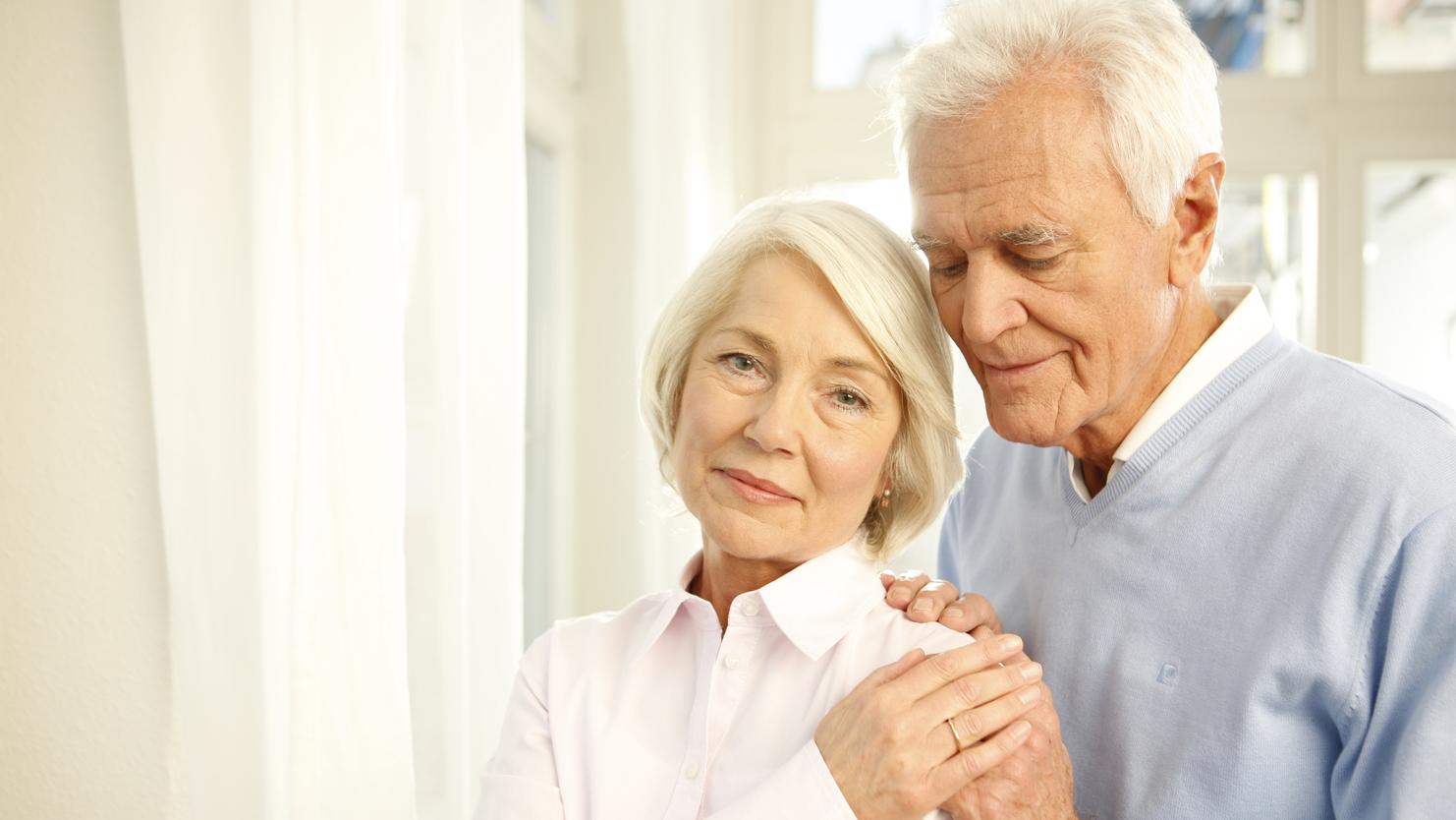 Ein älteres Paar steht nah beieinander, der Mann umarmt die Frau sanft, beide lächeln in eine ruhige Umgebung.